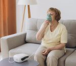 Sick senior woman has a viral infection or flu, labored breathing, asthma. An elderly female is using inhaler, nebulizer for self treatment sitting alone on the sofa at home